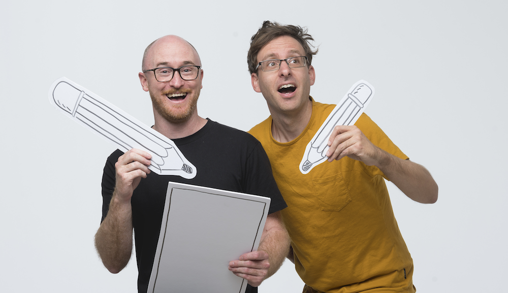 A photo of two white men (Ben Wood. and Andrew McDonald) with glasses, smiling. andholding giant, fake pencils. 